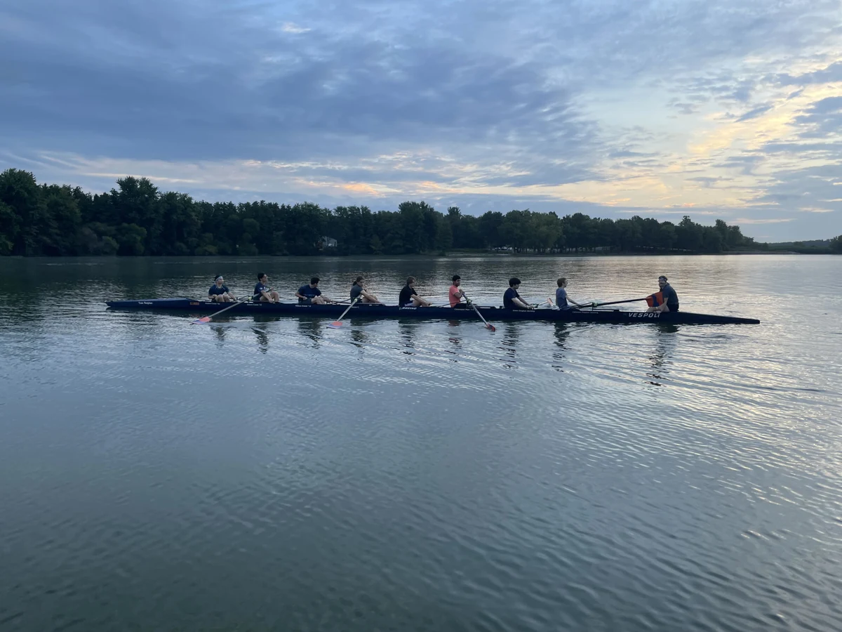 Rowing crew in a long shell on calm water beneath a cloudy sky